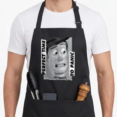 Woody the story of the apron - Grayscale Woody face flanked by two vertical white banners with black text — shown on a black cotton canvas apron against a white background