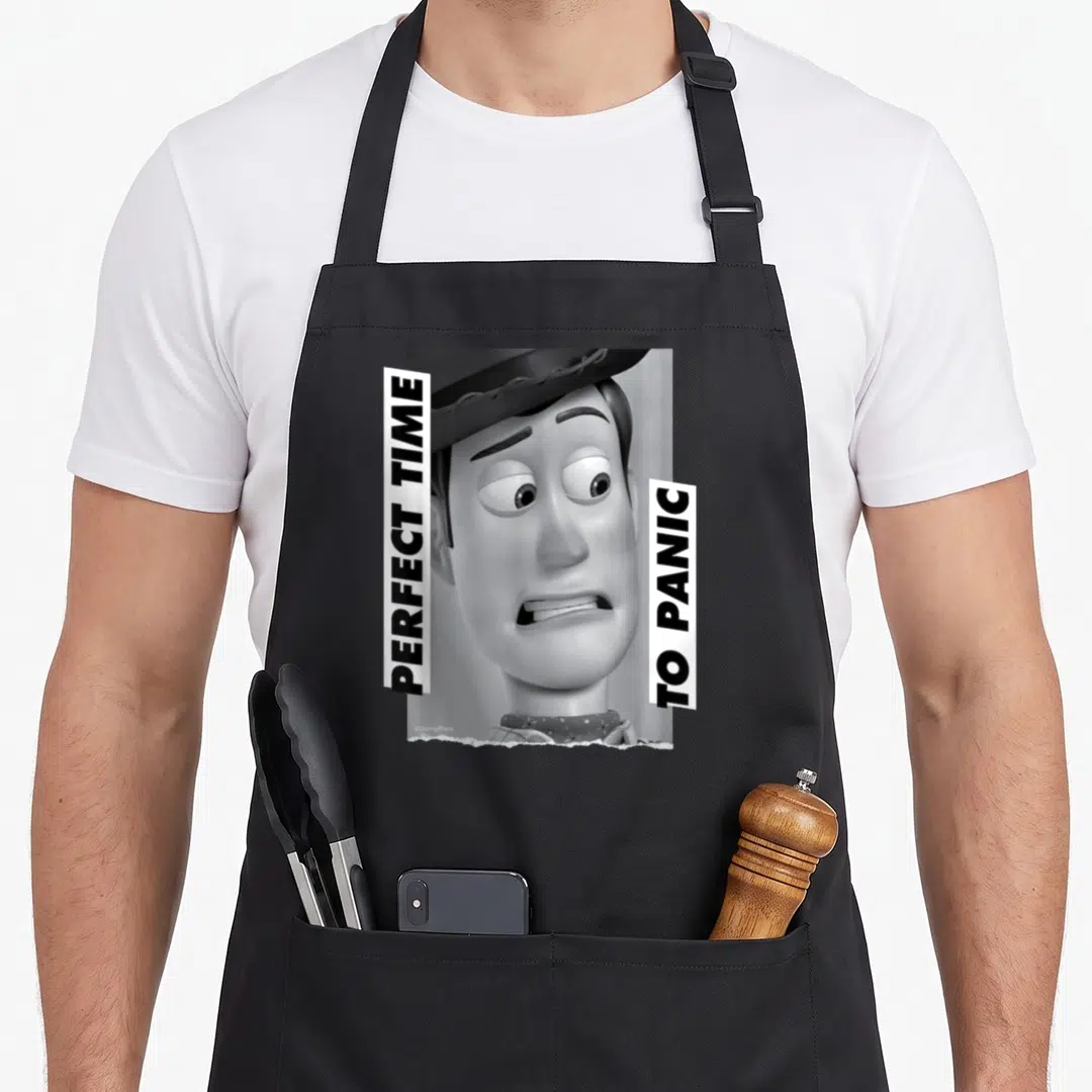 Woody the story of the apron - Grayscale Woody face flanked by two vertical white banners with black text — shown on a black cotton canvas apron against a white background