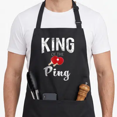 Ping king apron - KING OF THE Ping text with crossed red paddles and white ball in distressed style — shown on a black cotton canvas apron against a white background