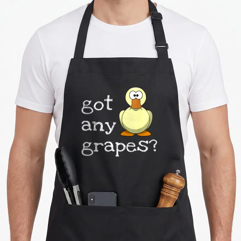 Retro grape duck apron - Yellow cartoon duck with orange beak next to stacked white text reading got any grapes — shown on a black cotton canvas apron against a white background