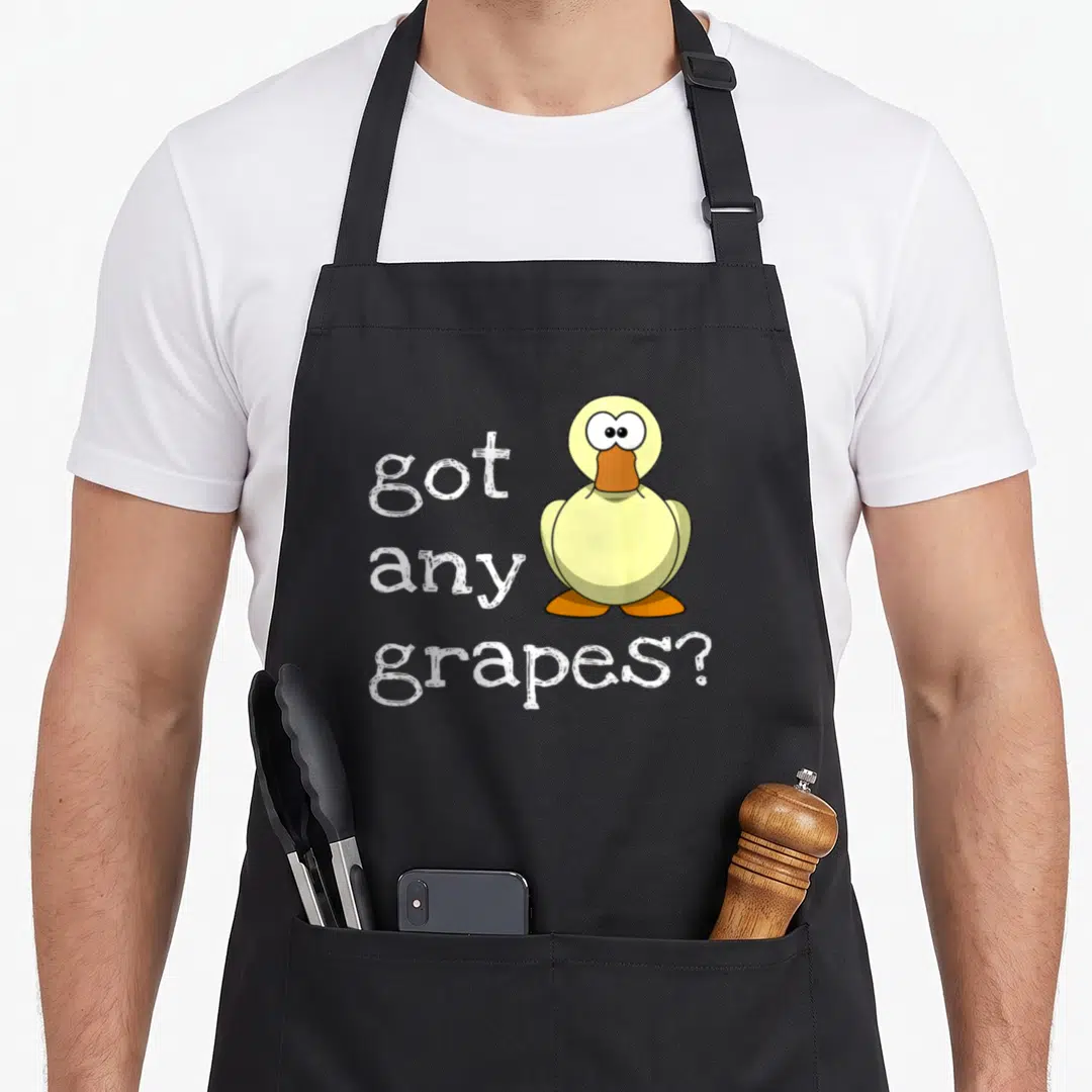 Retro grape duck apron - Yellow cartoon duck with orange beak next to stacked white text reading got any grapes — shown on a black cotton canvas apron against a white background