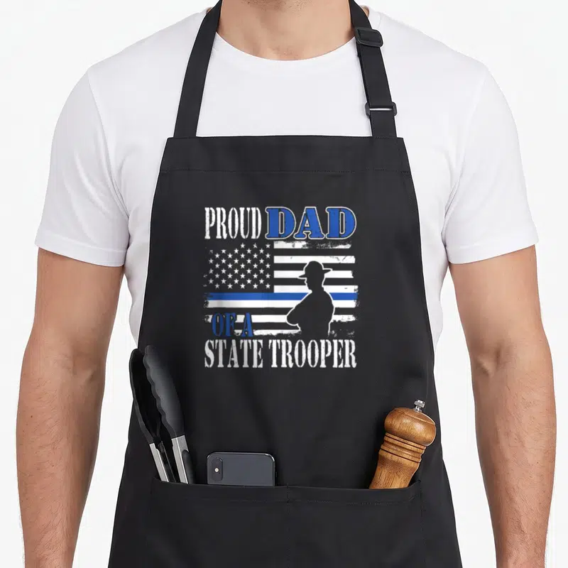 Funny proud police apron - Proud Dad of a State Trooper text over distressed flag with trooper silhouette — shown on a black cotton canvas apron against a white background