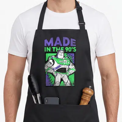 Retro 90s apron - Buzz Lightyear standing in green and purple frame below MADE IN THE 90'S text — shown on a black cotton canvas apron against a white background