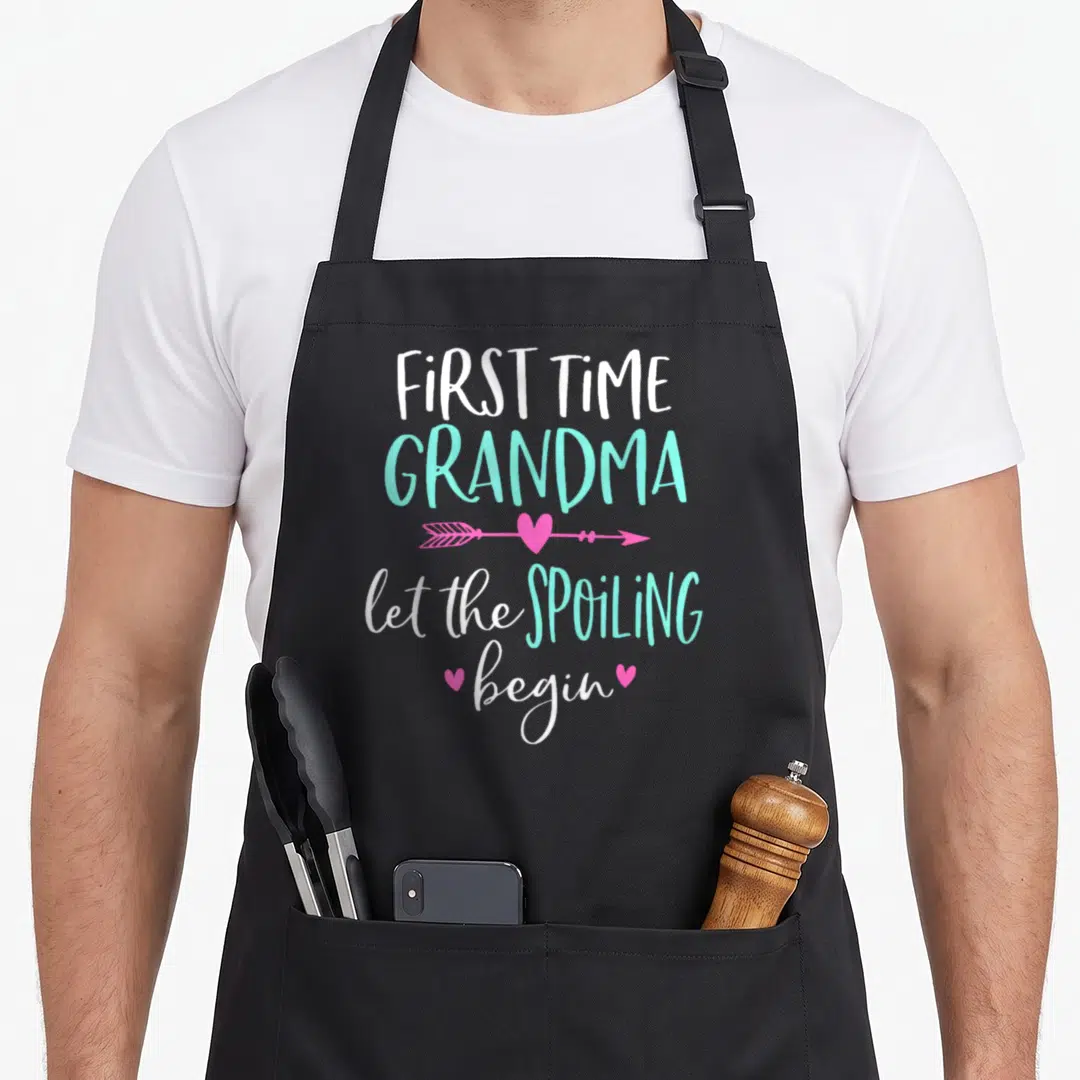 Spoiling grandma apron - Stacked text FIRST TIME GRANDMA with pink arrow and hearts in white turquoise pink — shown on a black cotton canvas apron against a white background