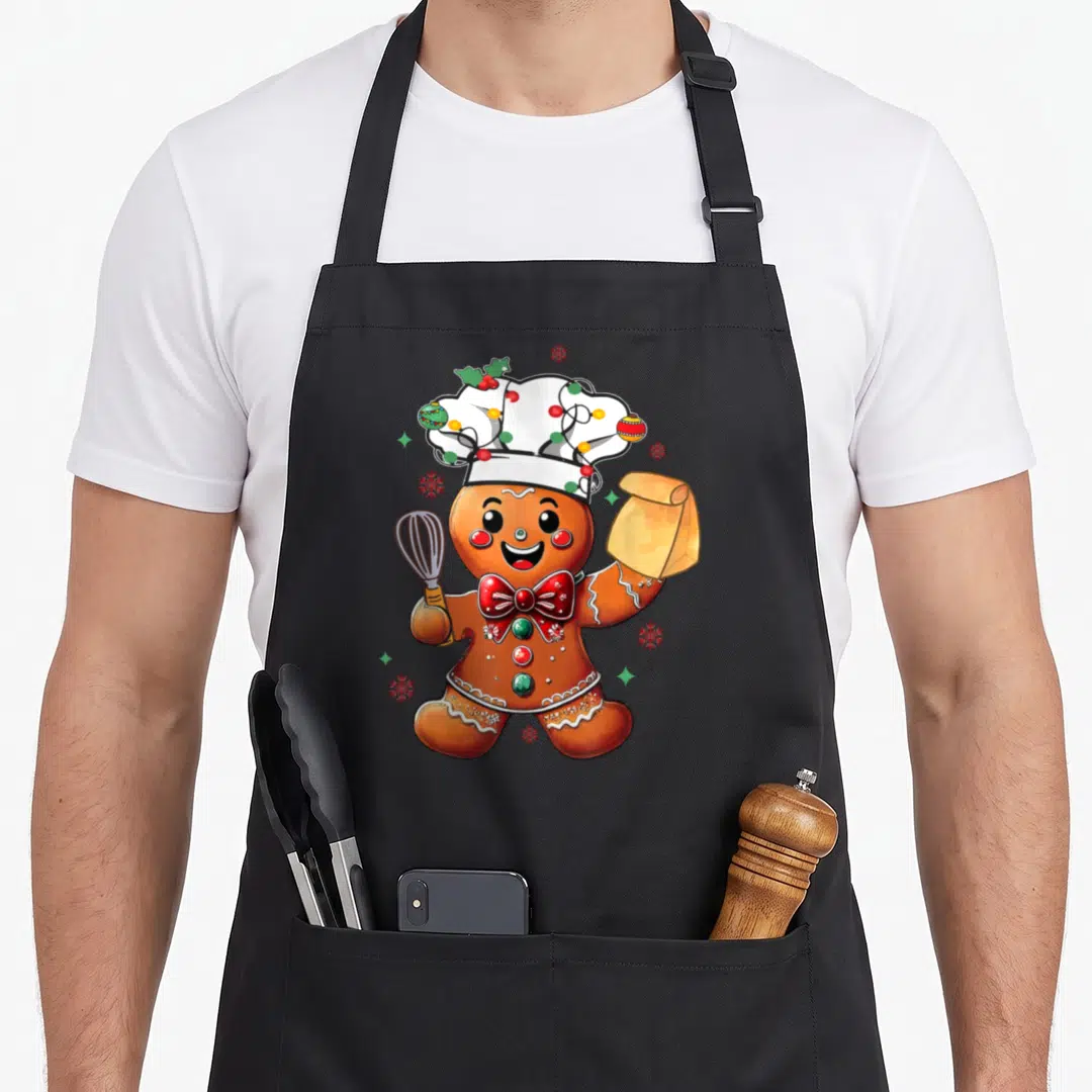 Baker women's christmas aprons - Cartoon gingerbread chef holding whisk and flour bag with Christmas hat — shown on a black cotton canvas apron against a white background