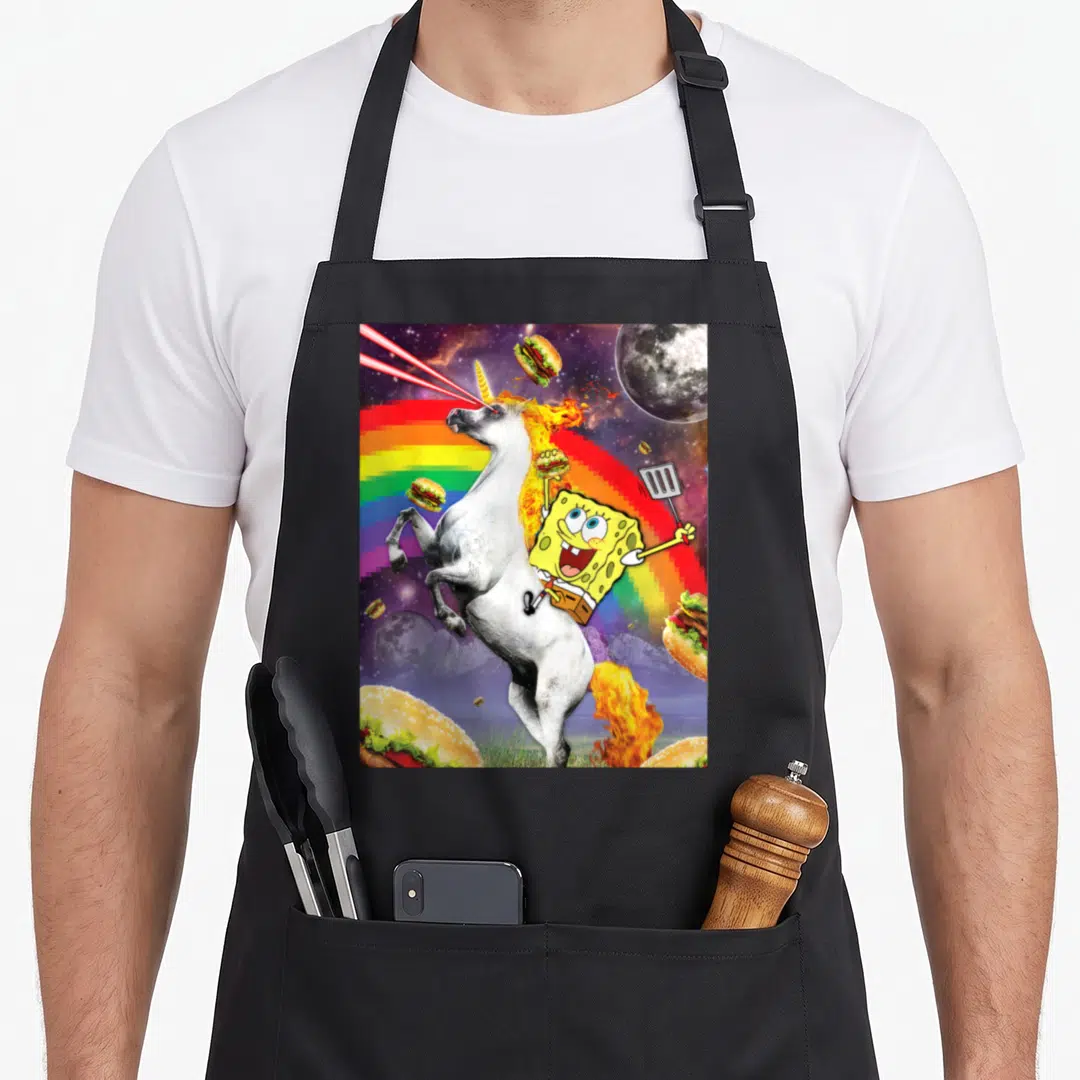 Squarepant rainbow apron - SpongeBob riding a rearing white unicorn with burgers and moons against a rainbow space — shown on a black cotton canvas apron against a white background