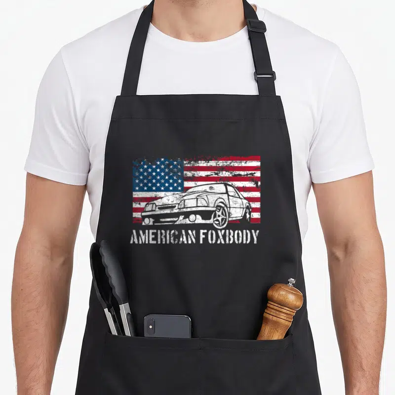 Foxbody muscle apron - Distressed white car on red white blue flag with AMERICAN FOXBODY text — shown on a black cotton canvas apron against a white background
