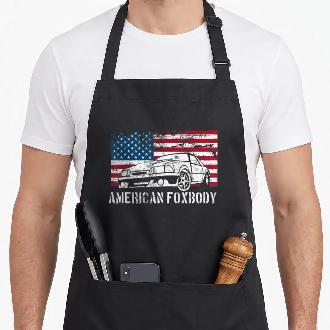 Foxbody muscle apron - Distressed white car on red white blue flag with AMERICAN FOXBODY text — shown on a black cotton canvas apron against a white background