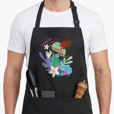 Come birthday apron - Mermaid with Birthday Wish Come True text amid pink, green, blue, purple aquatic plants — shown on a black cotton canvas apron against a white background
