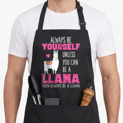 Llamas llama apron - Cartoon white llama with flower crown and text Always Be Yourself Unless You Can Be A Llama — shown on a black cotton canvas apron against a white background