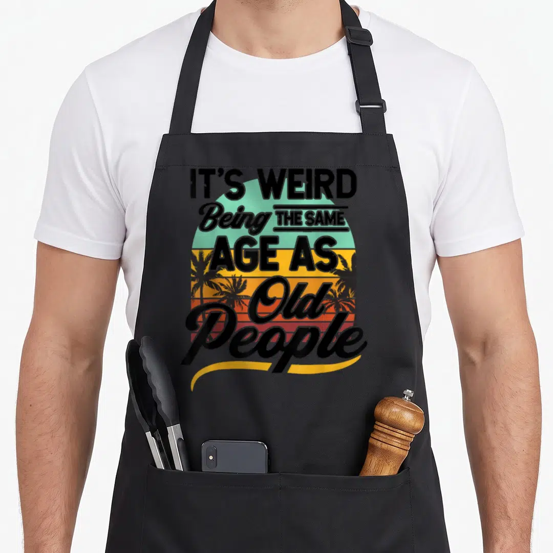Senior weird aprons - Retro sunset with palm trees and stacked black text reading IT'S WEIRD Being THE SAME AGE AS Old — shown on a black cotton canvas apron against a white background