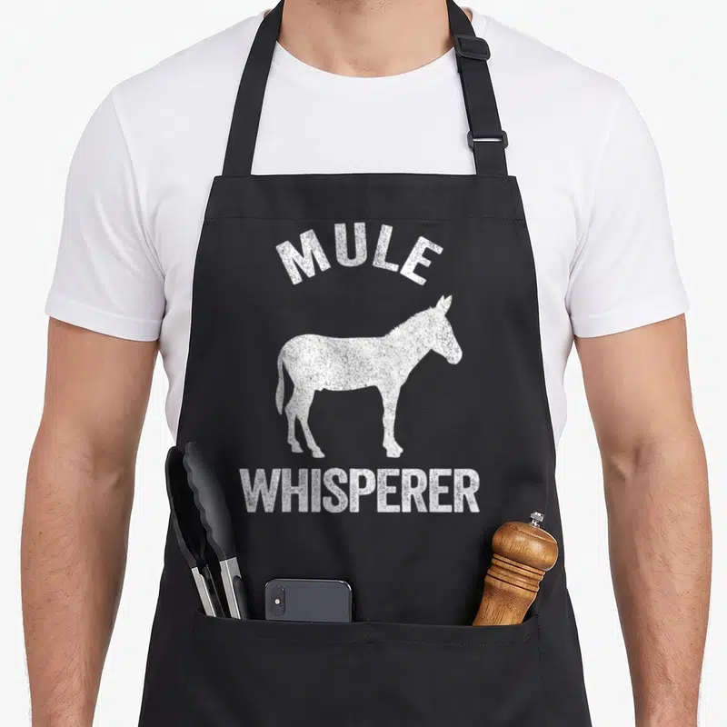 Mule whisperer funny apron - White distressed mule silhouette with arched MULE and straight WHISPERER text — shown on a black cotton canvas apron against a white background