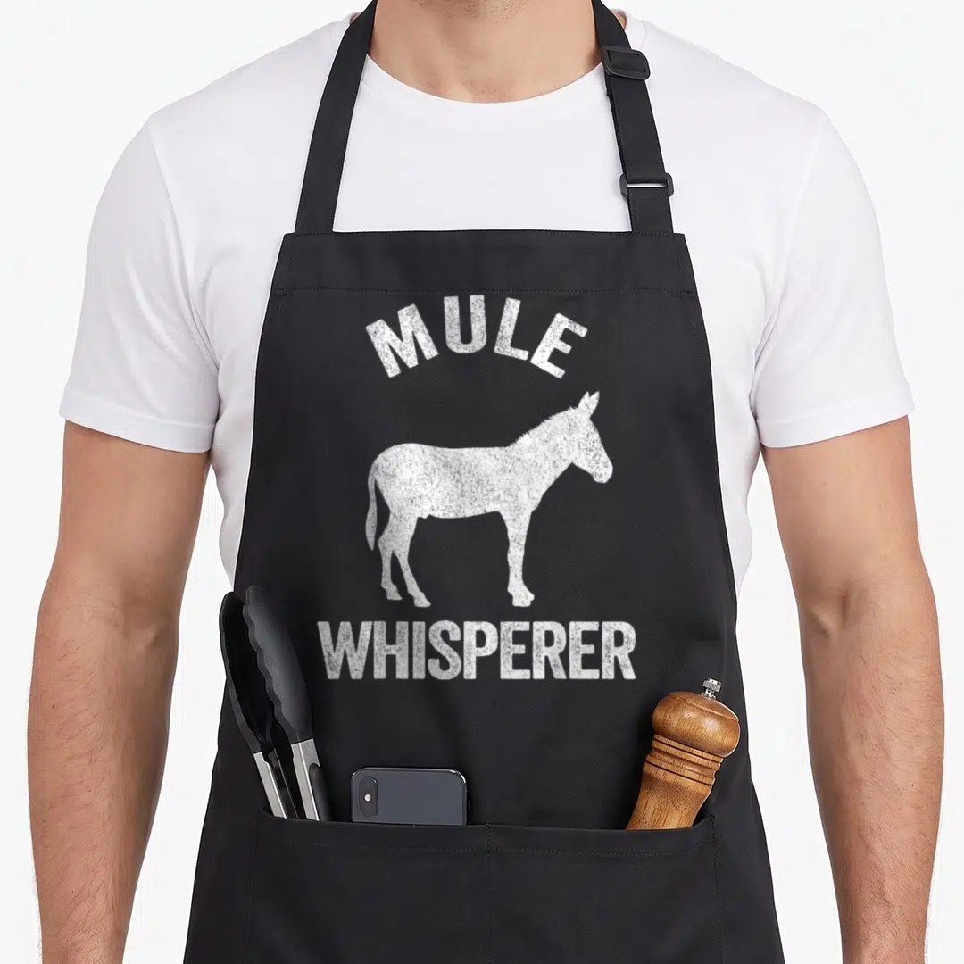 Mule whisperer funny apron - White distressed mule silhouette with arched MULE and straight WHISPERER text — shown on a black cotton canvas apron against a white background