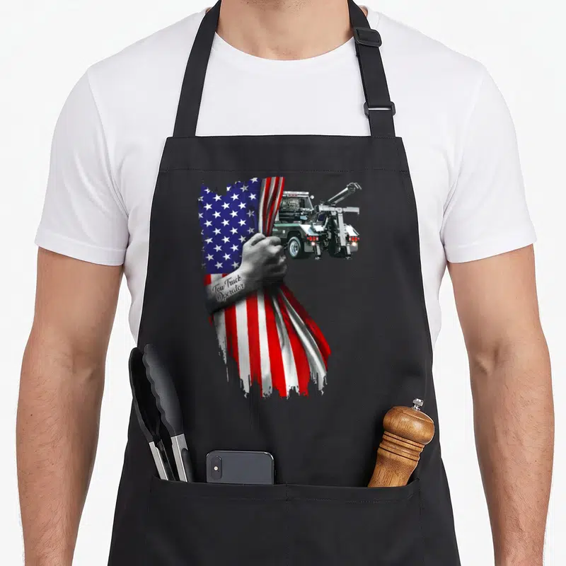 Tow truck american flag apron - Realistic hand holding draped American flag with tow truck behind it and text on wrist — shown on a black cotton canvas apron against a white background