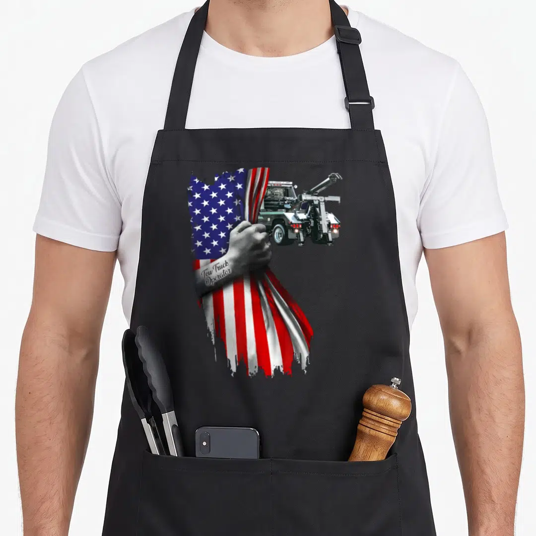 Tow truck american flag apron - Realistic hand holding draped American flag with tow truck behind it and text on wrist — shown on a black cotton canvas apron against a white background