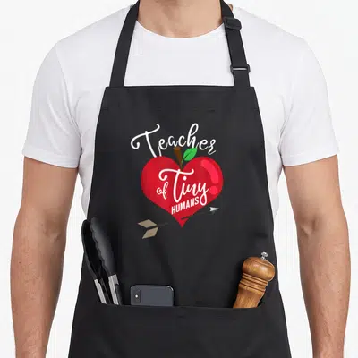 Human preschool teacher apron - Red heart apple with Teacher of Tiny HUMANS text and arrow piercing bottom — shown on a black cotton canvas apron against a white background