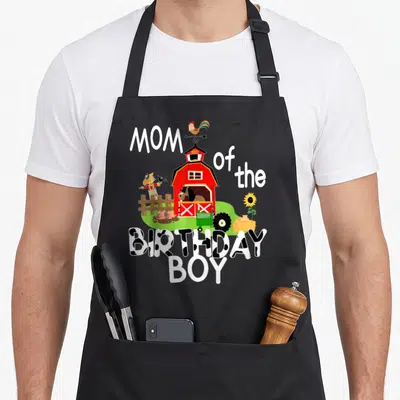Birthday farm aprons - Red barn with animals and cow-print text reading MOM of the BIRTHDAY BOY — shown on a black cotton canvas apron against a white background