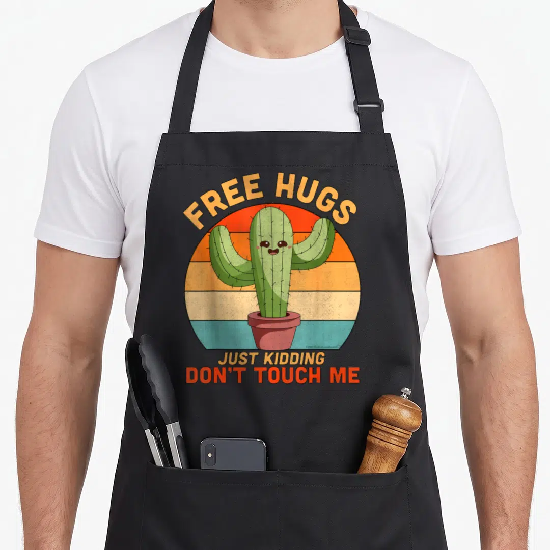 Hug cactus apron - Smiling green cactus in brown pot with striped background and text free hugs just kidding don't — shown on a black cotton canvas apron against a white background