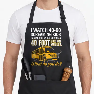 School wheel apron - I WATCH 40-60 SCREAMING KIDS text above yellow school bus illustration — shown on a black cotton canvas apron against a white background