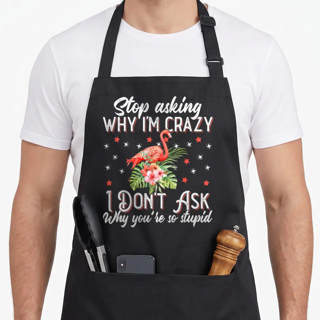 Asking flamingo apron - Sassy flamingo quote with hibiscus flowers and red stars on canvas — shown on a black cotton canvas apron against a white background