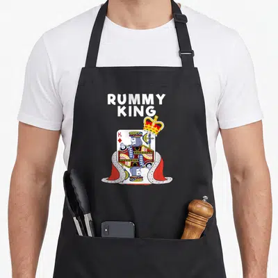 Rummy king apron - King of Diamonds playing card in ermine cape with floating gold crown and stacked white text — shown on a black cotton canvas apron against a white background
