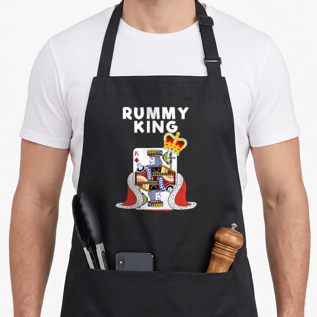 Rummy king apron - King of Diamonds playing card in ermine cape with floating gold crown and stacked white text — shown on a black cotton canvas apron against a white background