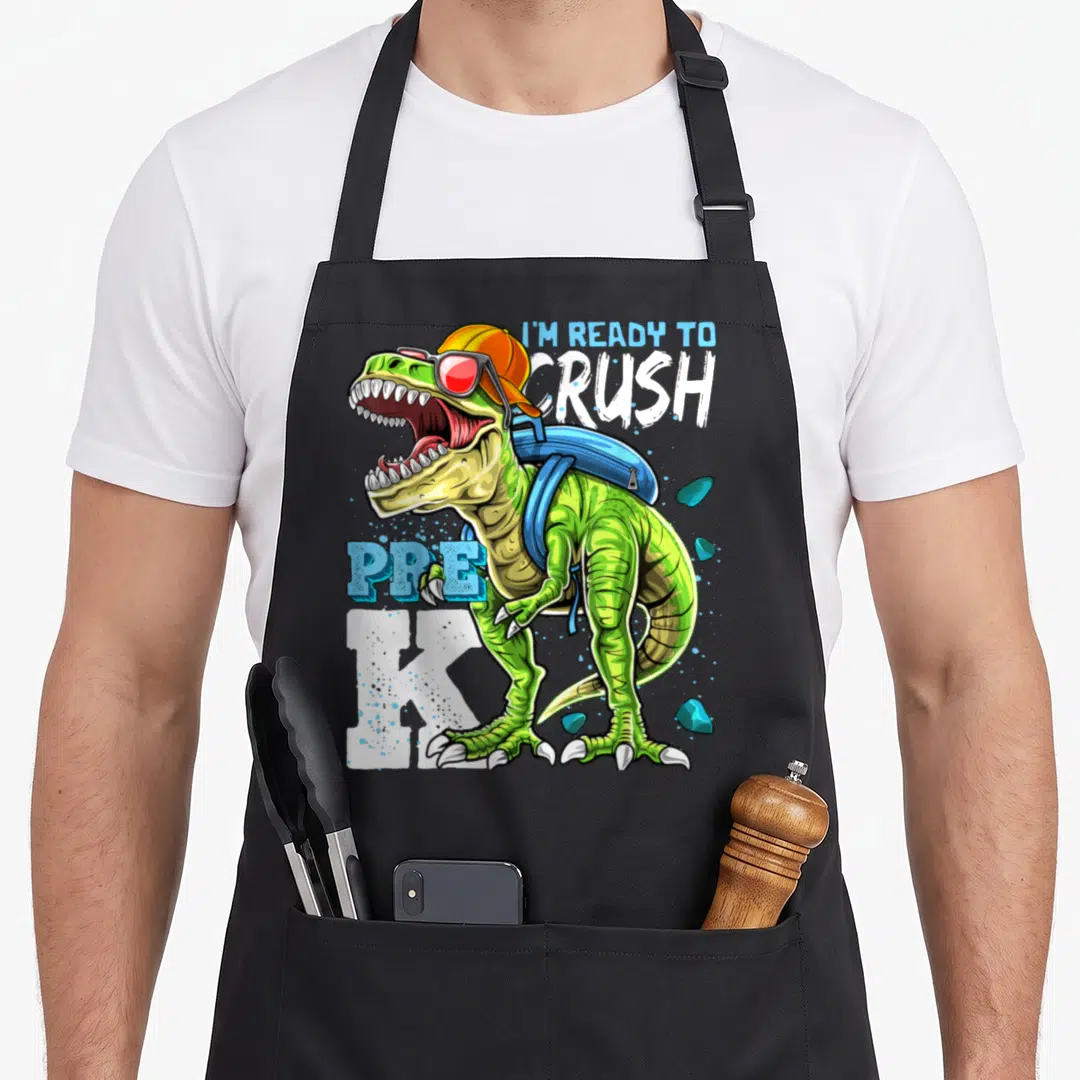 Pre dinosaur apron - Green T-Rex with orange cap, blue backpack, sunglasses, and I'M READY TO CRUSH PRE K text — shown on a black cotton canvas apron against a white background