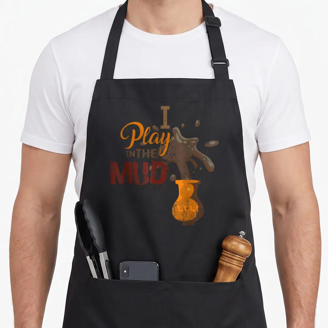 Play mud guy apron - Stacked I Play IN THE MUD text beside a brown mud splash from an orange vase in distressed orange — shown on a black cotton canvas apron against a white background