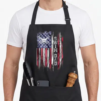 Flag swimming dad apron - Distressed US flag with white swimmer icon and vertical SWIM DAD text in red white blue — shown on a black cotton canvas apron against a white background