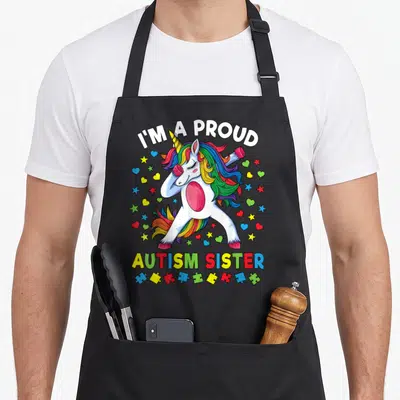 Proud unicorn apron - Dabbing unicorn with rainbow mane, puzzle pieces, and I'M A PROUD, AUTISM SISTER text — shown on a black cotton canvas apron against a white background
