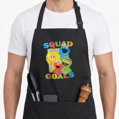 Squad street apron - Four Sesame Street characters clustered between arched SQUAD GOALS text in yellow, blue, red — shown on a black cotton canvas apron against a white background