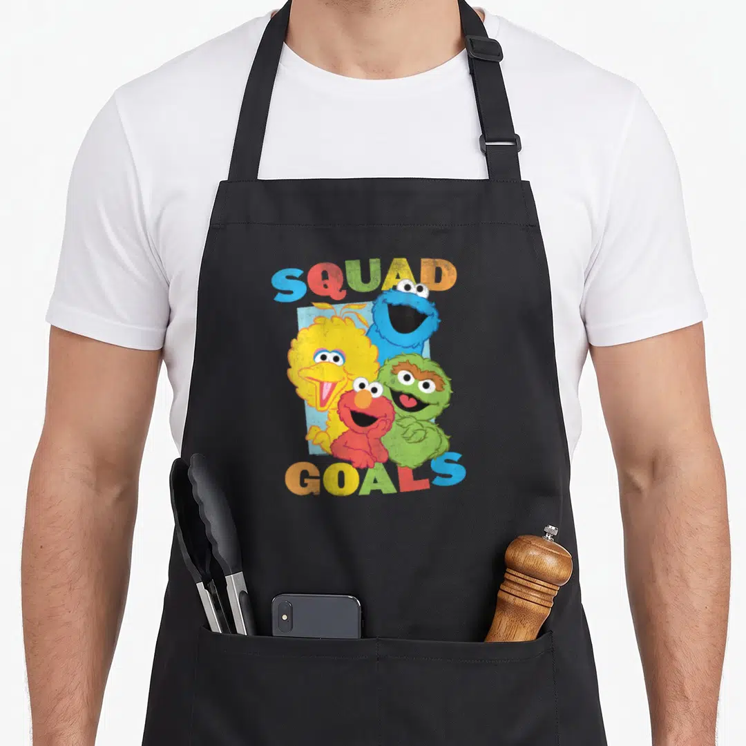 Squad street apron - Four Sesame Street characters clustered between arched SQUAD GOALS text in yellow, blue, red — shown on a black cotton canvas apron against a white background