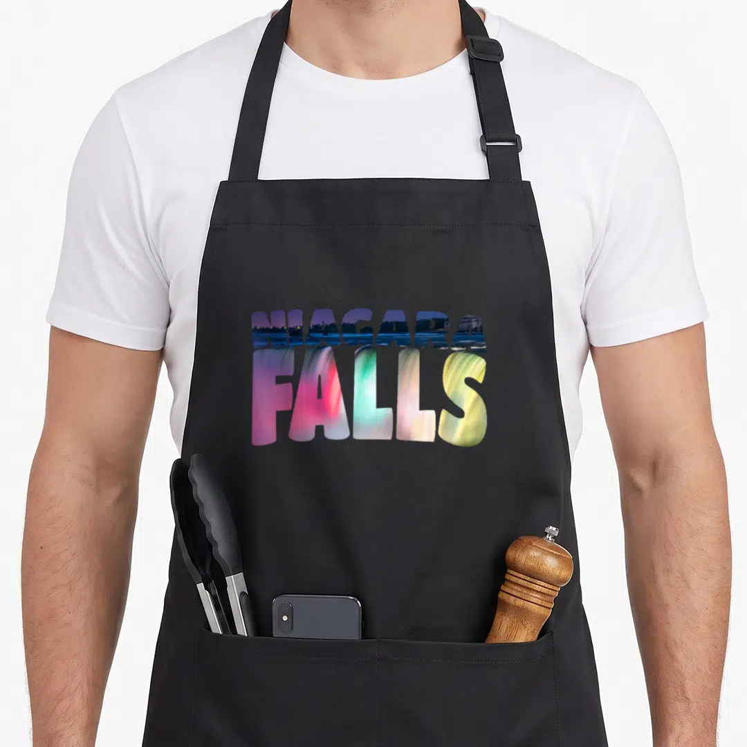 Night fall apron - Stacked NIAGARA FALLS text filled with blue pink green and yellow scenic imagery — shown on a black cotton canvas apron against a white background