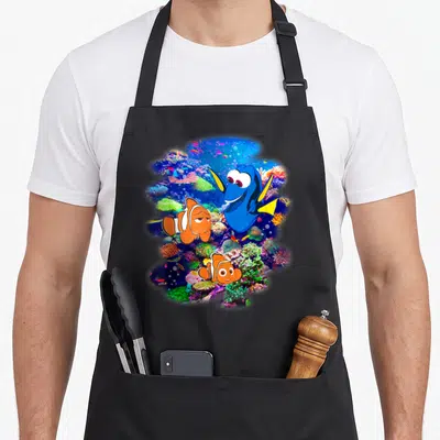 Finding rainbow apron - Three cartoon fish on blue, orange, pink, and green coral reef background — shown on a black cotton canvas apron against a white background