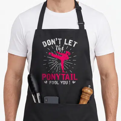 Karate fool apron - Pink martial artist kicking with text DON'T LET the 2 PONYTAIL FOOL YOU ! and two hearts — shown on a black cotton canvas apron against a white background