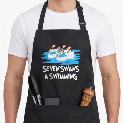 Seven swan christmas apron - Seven cartoon swans in red hats swimming in blue water with white text below — shown on a black cotton canvas apron against a white background