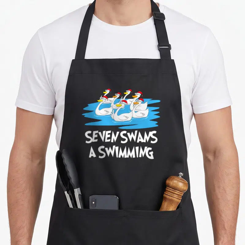 Seven swan christmas apron - Seven cartoon swans in red hats swimming in blue water with white text below — shown on a black cotton canvas apron against a white background