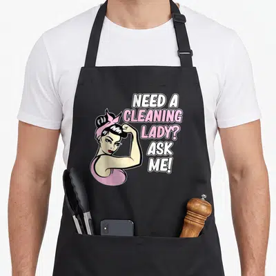 Retro housekeeping housekeeper apron - Cartoon woman flexing arm next to NEED A CLEANING LADY ASK ME text in pink — shown on a black cotton canvas apron against a white background