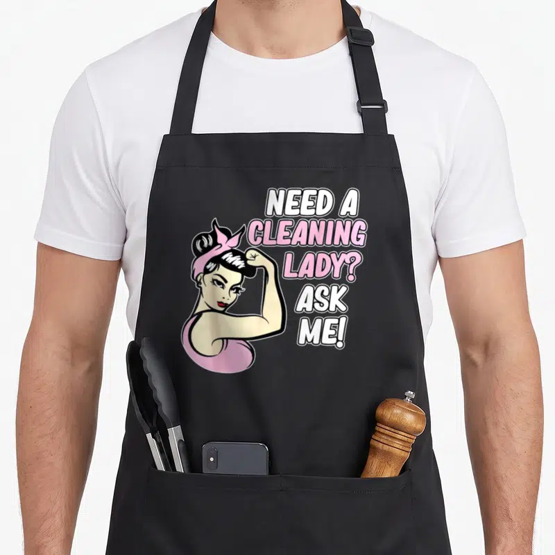 Retro housekeeping housekeeper apron - Cartoon woman flexing arm next to NEED A CLEANING LADY ASK ME text in pink — shown on a black cotton canvas apron against a white background