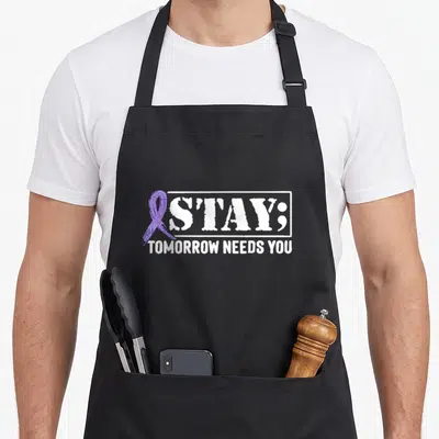 Stay suicide prevention apron - Purple awareness ribbon next to distressed white S'TAY; text and TOMORROW NEEDS YOU — shown on a black cotton canvas apron against a white background