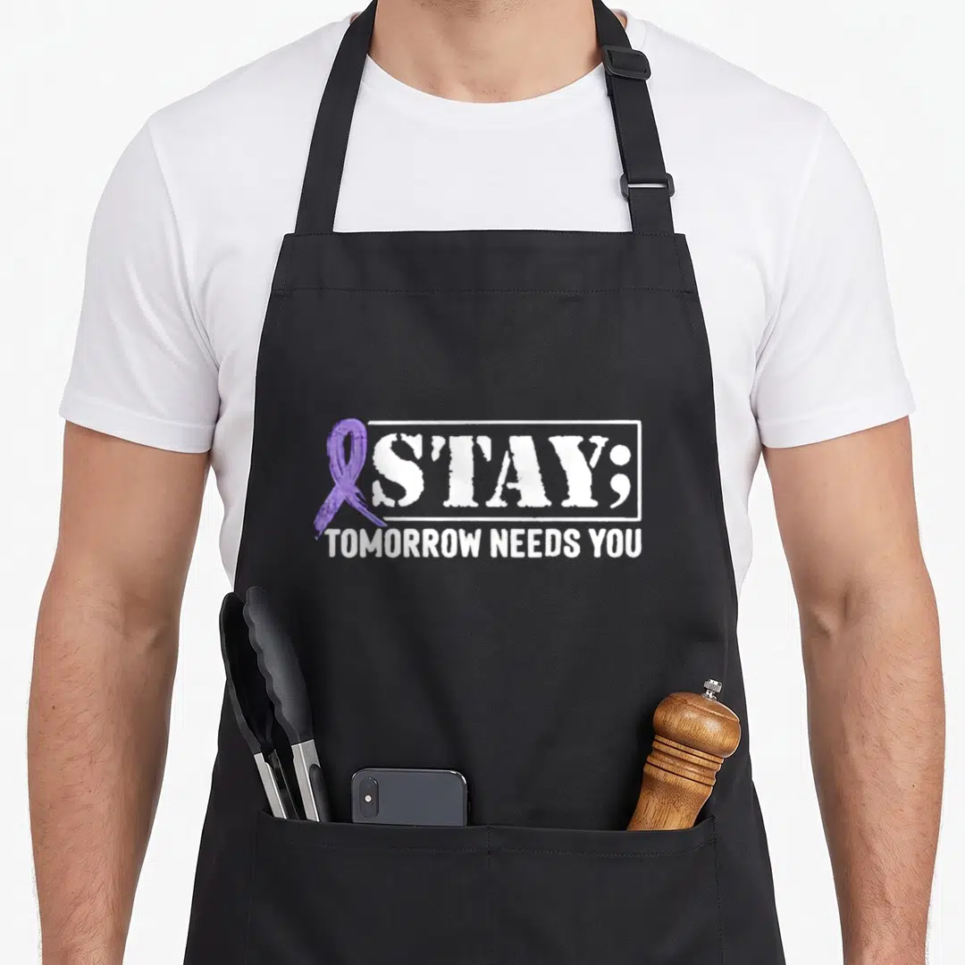 Stay suicide prevention apron - Purple awareness ribbon next to distressed white S'TAY; text and TOMORROW NEEDS YOU — shown on a black cotton canvas apron against a white background
