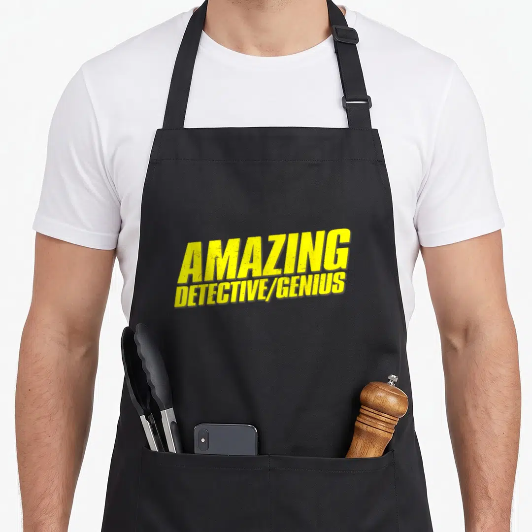 Amazing halloween apron - Yellow stacked text reading AMAZING DETECTIVE/GENIUS on a dark background — shown on a black cotton canvas apron against a white background