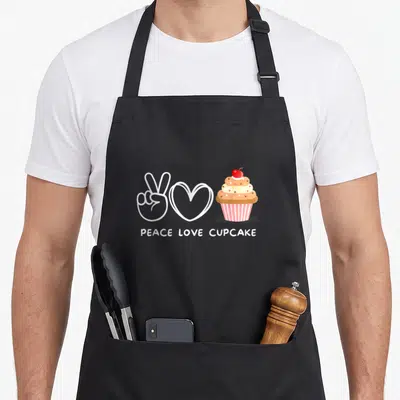 Peace cupcake apron - Cartoon peace sign, white heart, and pink striped cupcake with red cherry and sprinkles — shown on a black cotton canvas apron against a white background