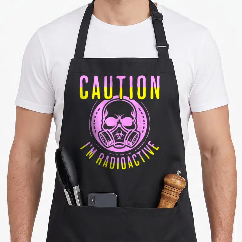 Warrior radiation aprons - Pink and yellow Caution I'm Radioactive text with skull gas mask cartoon — shown on a black cotton canvas apron against a white background