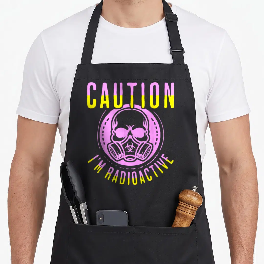 Warrior radiation aprons - Pink and yellow Caution I'm Radioactive text with skull gas mask cartoon — shown on a black cotton canvas apron against a white background