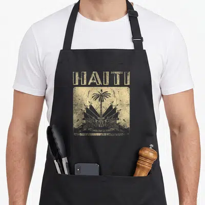 Haitian haiti apron - Distressed tan square with HAITI text, palm tree, cannons, and motto L'UNION FAIT LA FORCE — shown on a black cotton canvas apron against a white background
