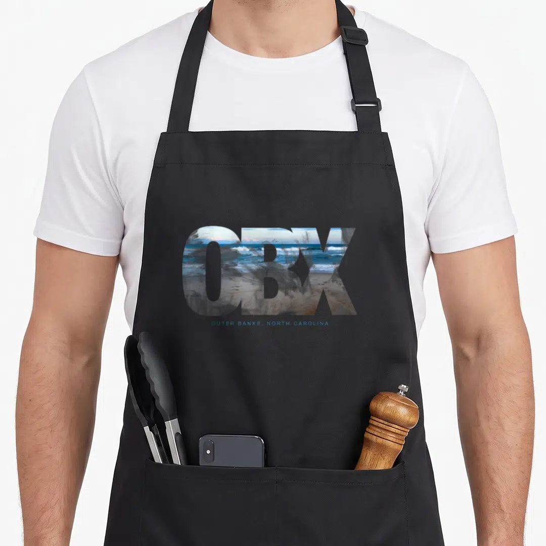 Rja outer banks apron - Large OBX letters filled with ocean waves and sand, with smaller blue text below — shown on a black cotton canvas apron against a white background