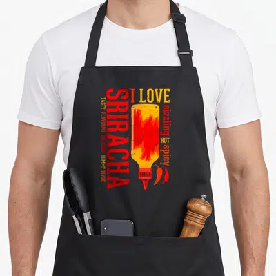 Sauce sriracha apron - Distressed red sriracha bottle with fire and stacked adjectives like YUMMY and DIVINE beside two — shown on a black cotton canvas apron against a white background