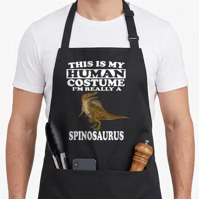 Human dinosaur apron - Cartoon Spinosaurus flanked by white text saying This Is My Human Costume I'm Really — shown on a black cotton canvas apron against a white background