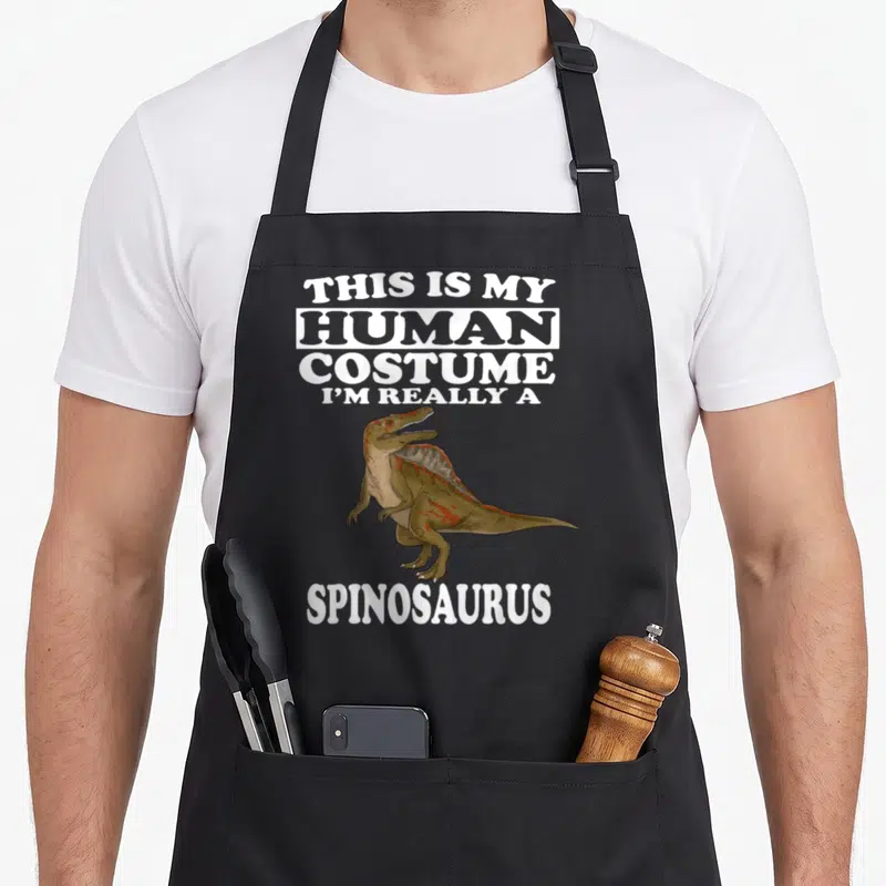 Human dinosaur apron - Cartoon Spinosaurus flanked by white text saying This Is My Human Costume I'm Really — shown on a black cotton canvas apron against a white background